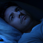 Pessoa com expressão de preocupação e olhos abertos na cama no escuro, ilustrando o sono ansioso e a mente acelerada.